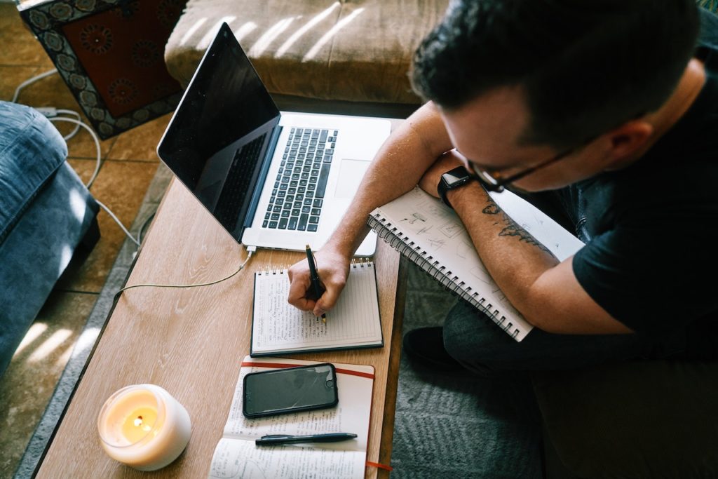 man shown from above writes in a notebook with a laptop on table as he works remotely from home